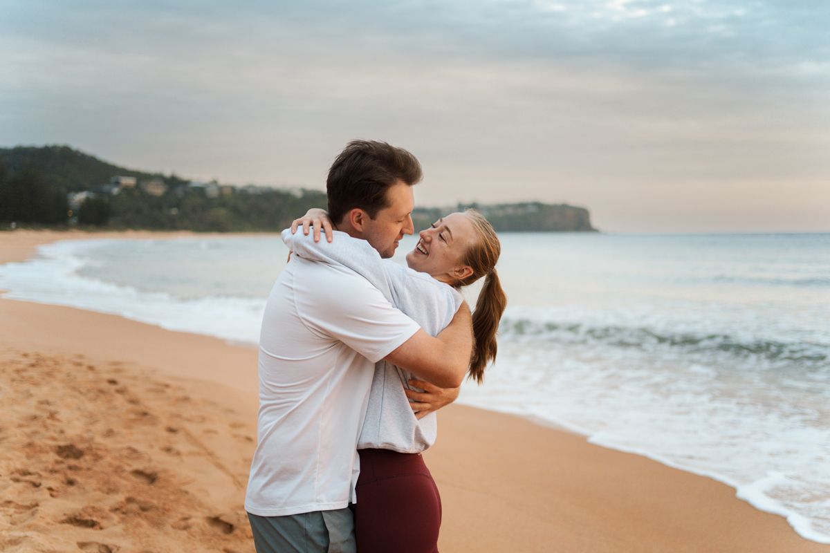 Couple hugging and looking at each other on a sandy Newport beach at sunrise with ocean waves in the background during surprise engagement