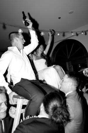 Wedding couple being lifted on chairs during traditional celebratory dance at reception.