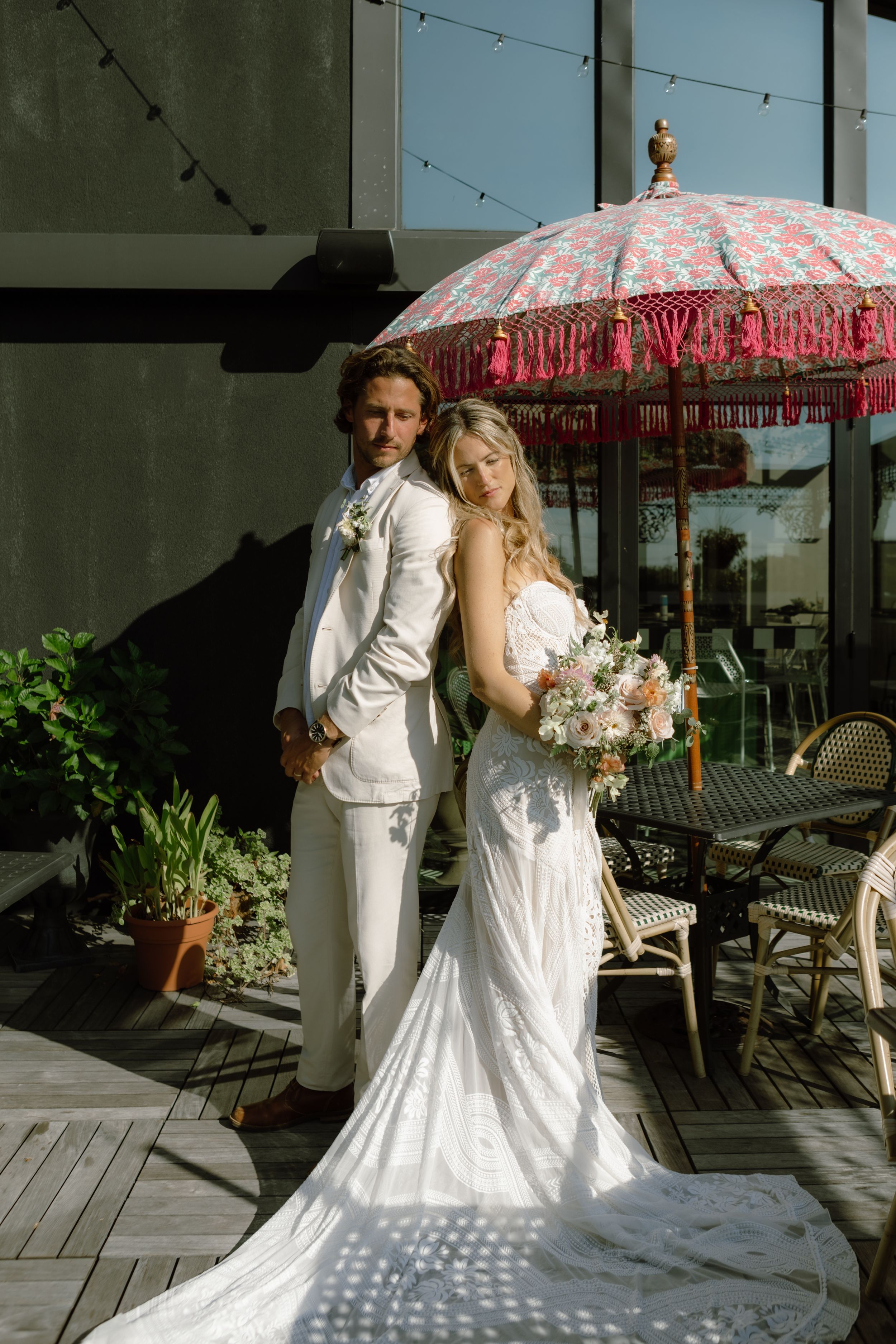A wedding couple poses elegantly under a pink fringed parasol on an outdoor deck.