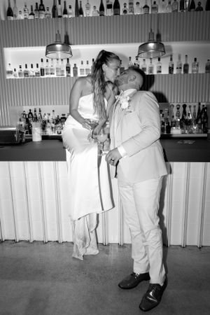 A romantic couple shares a kiss at a bar with bottles displayed in the background.