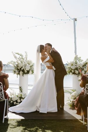 Romantic first kiss during outdoor wedding ceremony under string lights and white floral arch.
