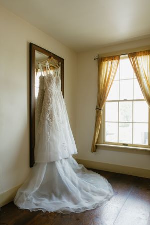 White lace wedding dress hanging by window with flowing train and sheer yellow curtains.