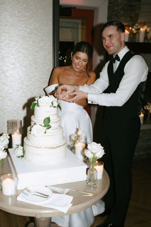 A couple cuts their elegant white tiered wedding cake decorated with roses.