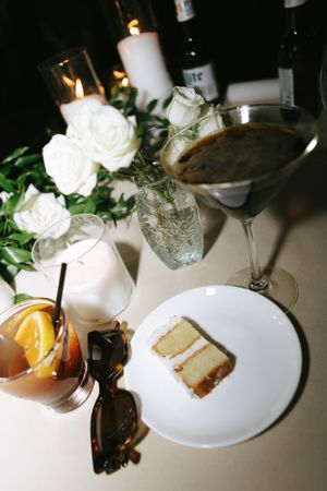 Wedding reception table setting with cocktails, white roses, and a slice of cake.