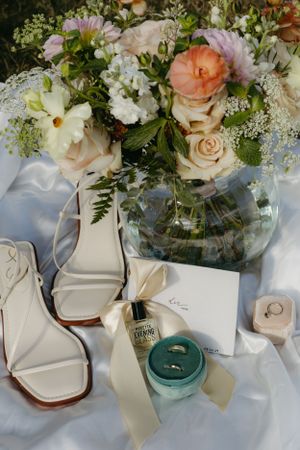 Wedding detail shot of floral arrangement, bridal shoes, and ring boxes on white fabric.