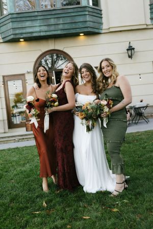 Four women in jewel-toned dresses laugh together while holding bouquets on a lawn.