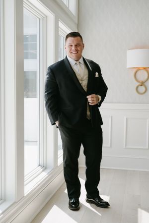 Groom in classic black tuxedo poses by window in elegant wedding venue.