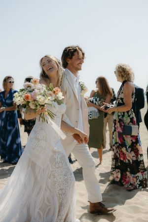 A wedding couple laughs joyfully while walking on a sunny beach with guests in the background.
