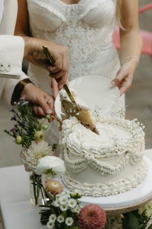 Hands cutting into an elegant white wedding cake decorated with intricate lace patterns.