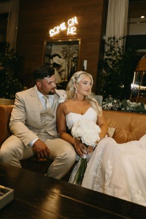 Wedding couple resting on tan leather couch under neon signage in evening reception setting.