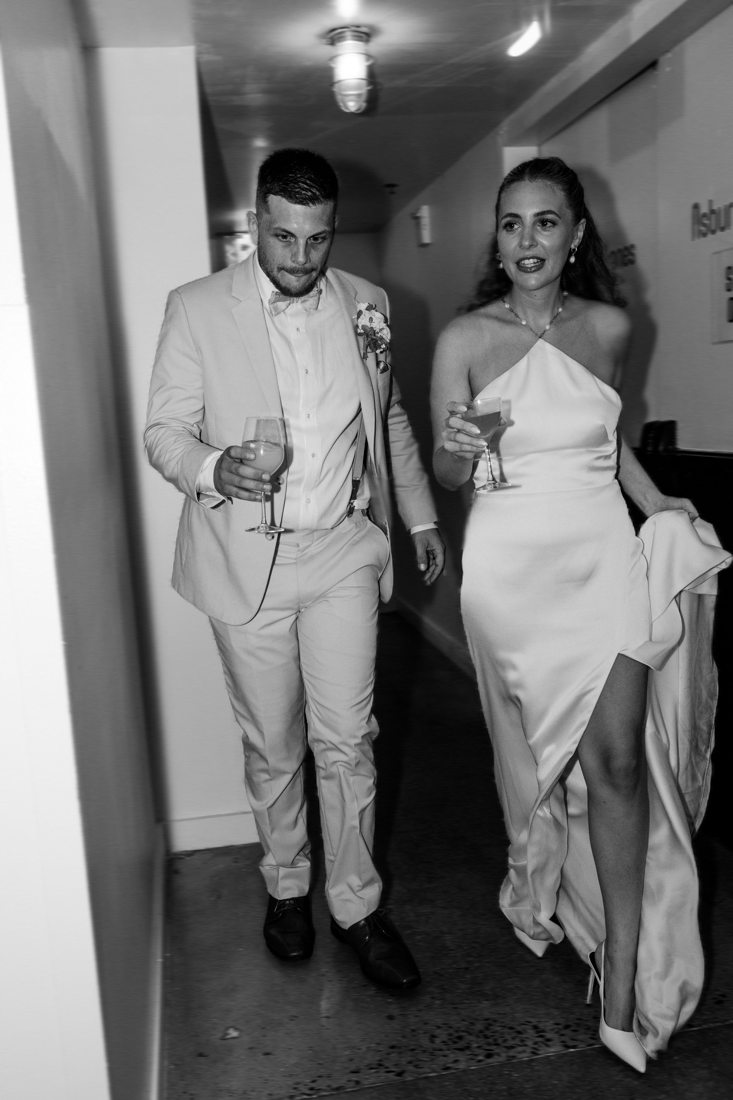 A couple in matching light-colored attire walks down a hallway holding drinks.