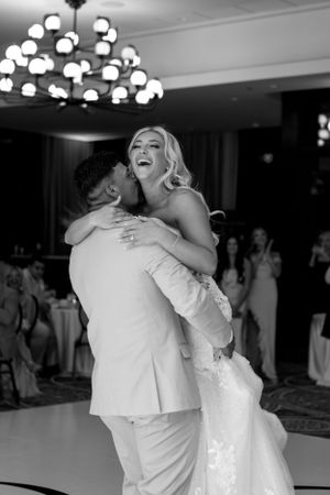 Black and white photo of a couple sharing a joyful dance moment at their wedding reception under chandelier lighting.