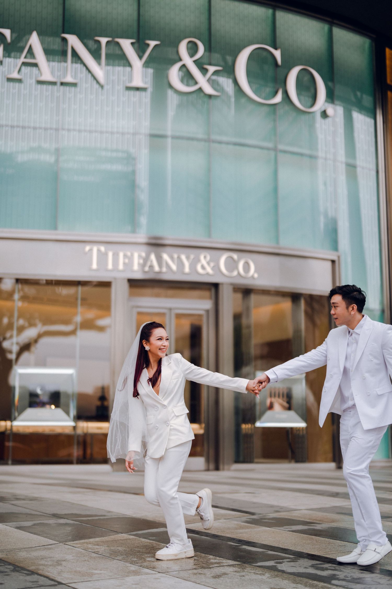 Couple in white outfits posing playfully outside Tiffany & Co store.