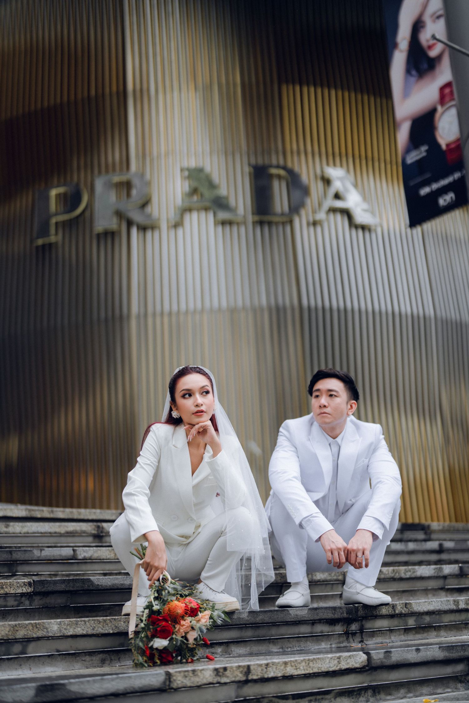 Two people in white attire sitting on concrete steps with a bouquet.