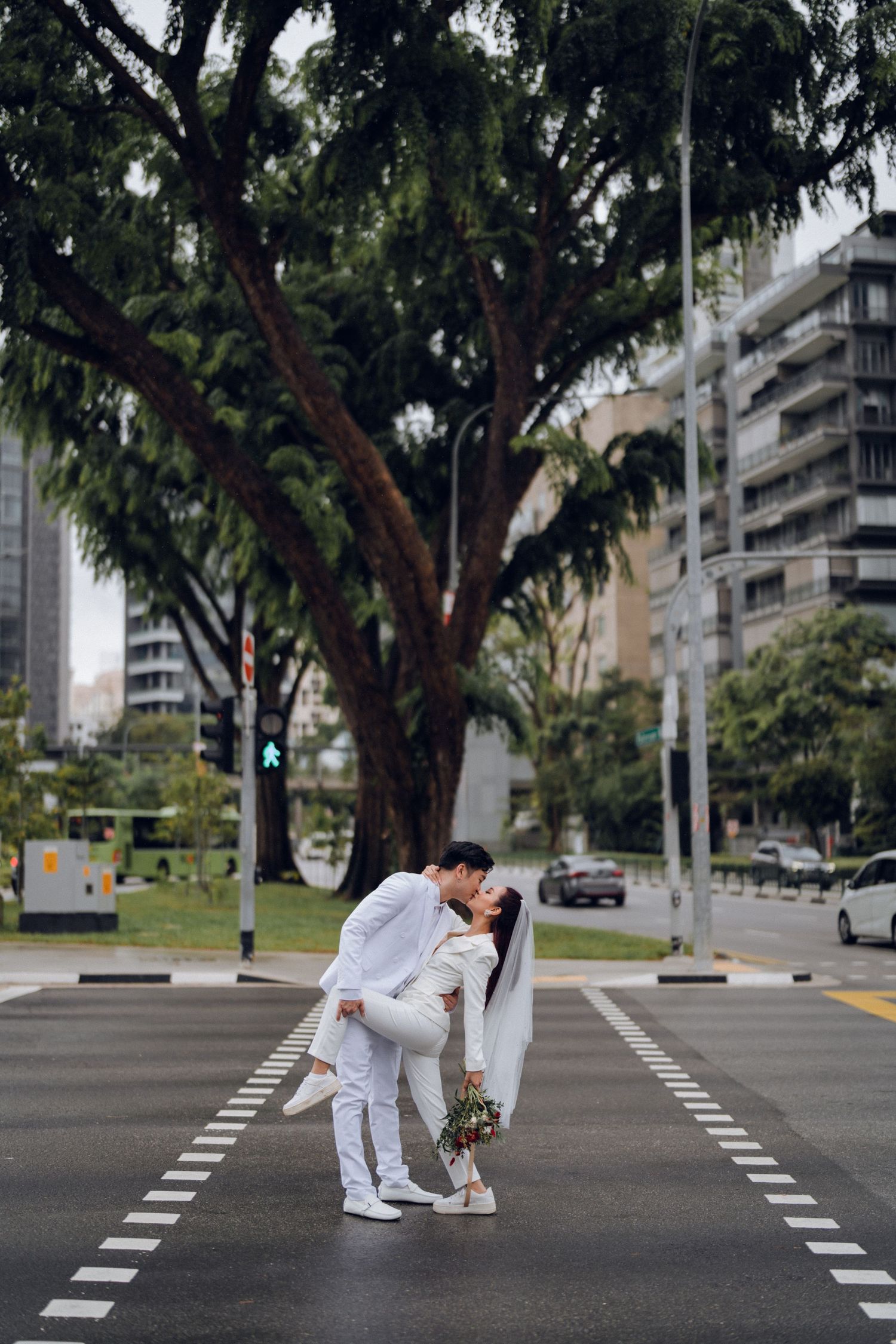 A couple in white outfits posing playfully on a crosswalk with trees in the background.