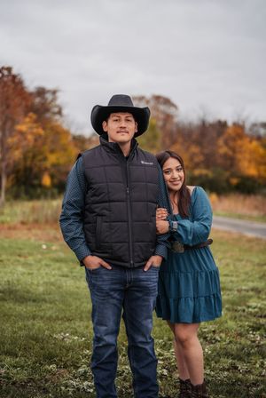 A couple poses together outdoors on a grassy hill with autumn foliage in the background.