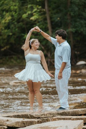Couple in white outfits dancing on rocks by a creek, man spinning woman in a white dress outdoors.