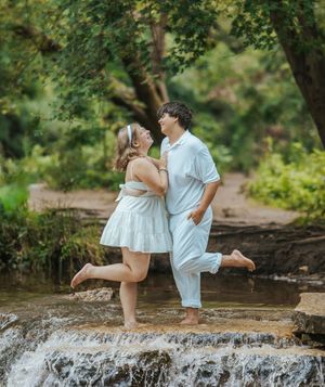 Couple in white outfits standing barefoot on a rocky waterfall surrounded by lush green trees.