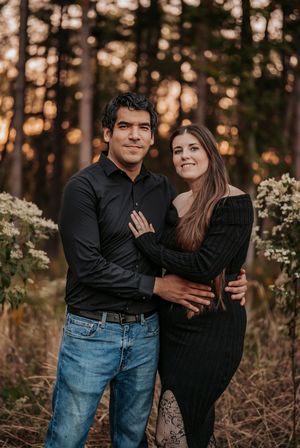 A couple embraces in a romantic forest setting at sunset, wearing black tops against a backdrop of wildflowers and trees.