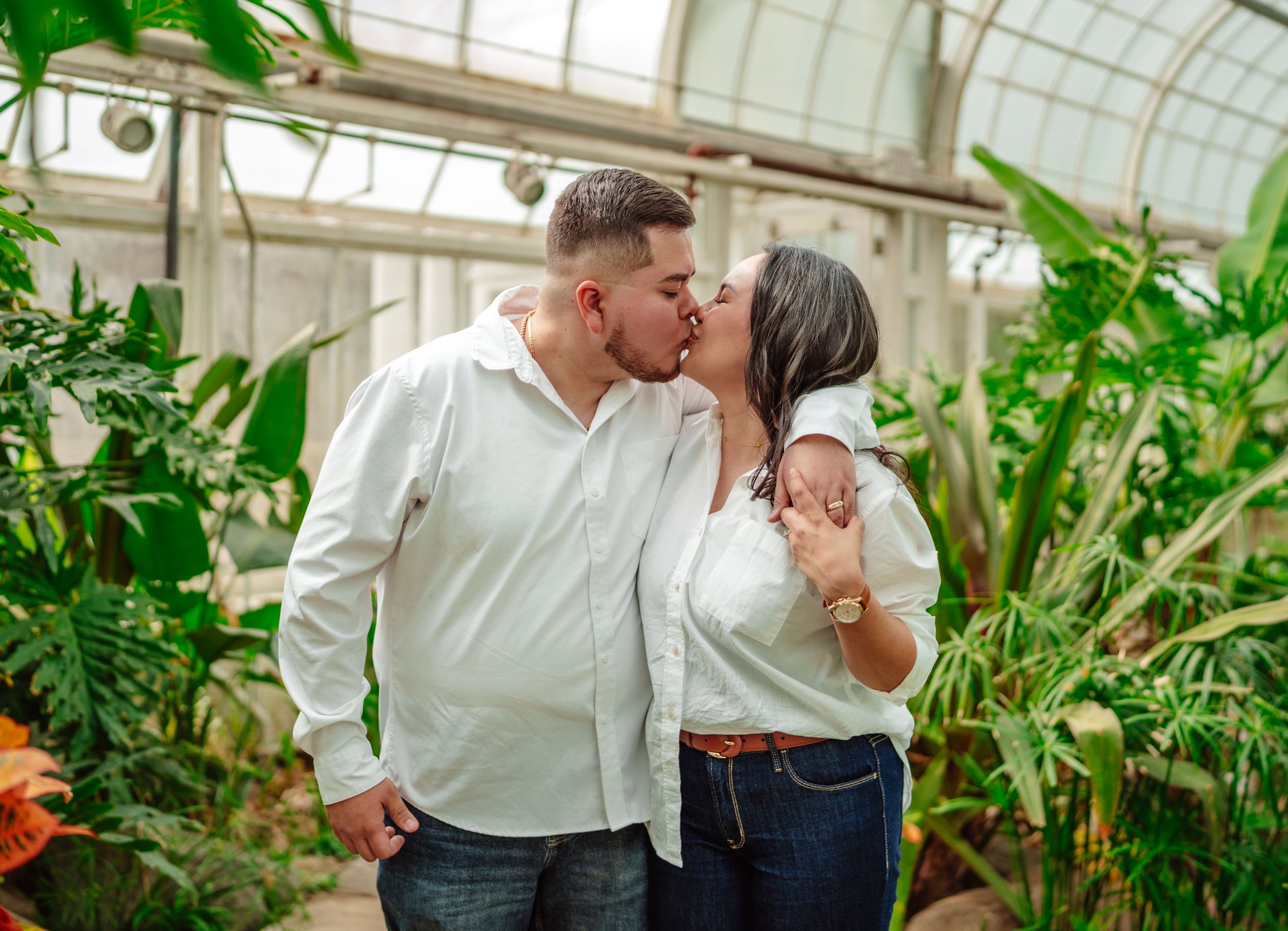 A couple shares a romantic moment surrounded by lush greenery in a sunlit greenhouse conservatory.