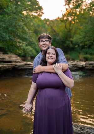 A couple shares a tender embrace while standing by a peaceful river at sunset in matching purple attire.