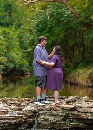 A couple stands together on a rocky creek bed surrounded by lush green forest scenery.