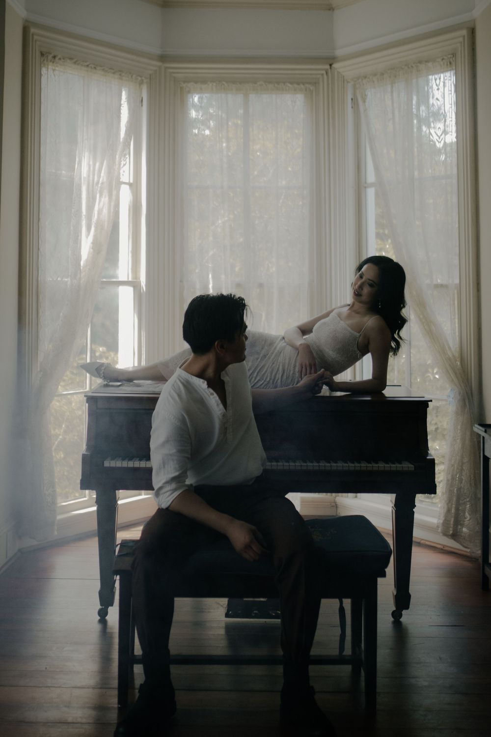 Moody scene of two people at a piano in a sunlit room with sheer curtains.