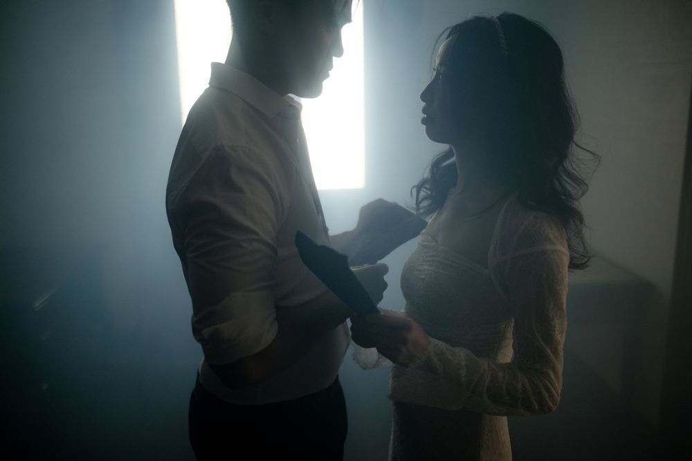 Two figures stand in silhouette against bright window light in a moody setting.
