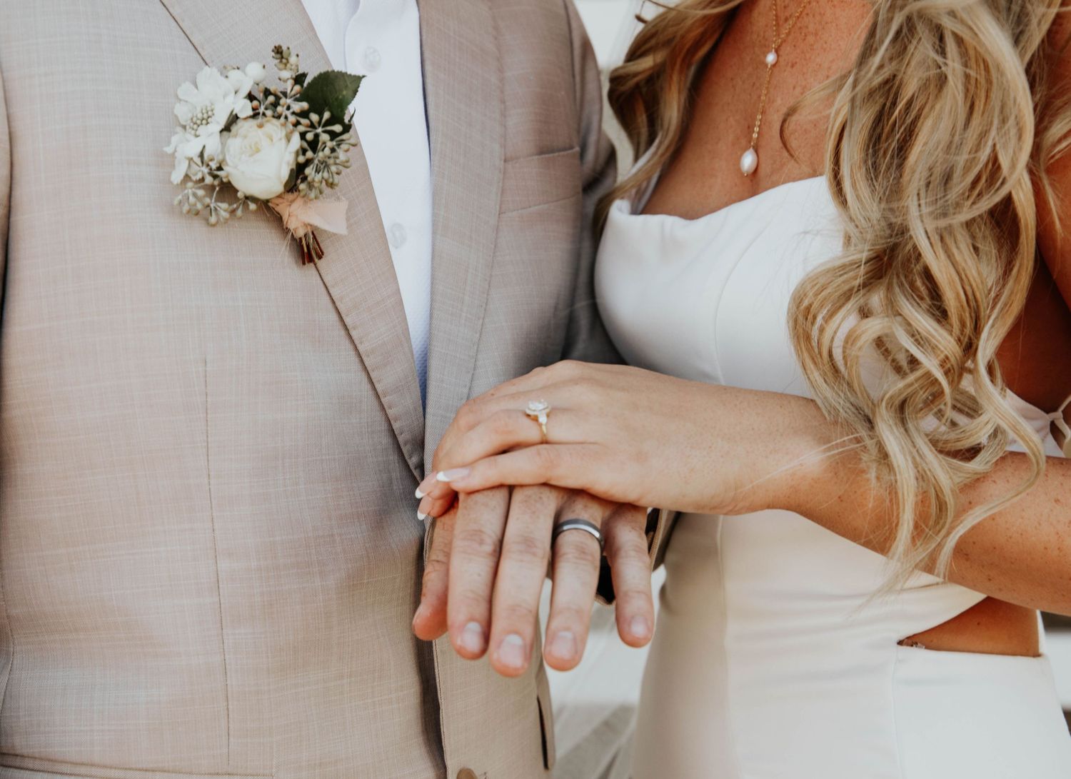 Close-up details of wedding attire showing hands, rings, and dress fabric in neutral tones.