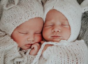 Two sleeping newborn babies in white knit hats lying together on soft white fabric.