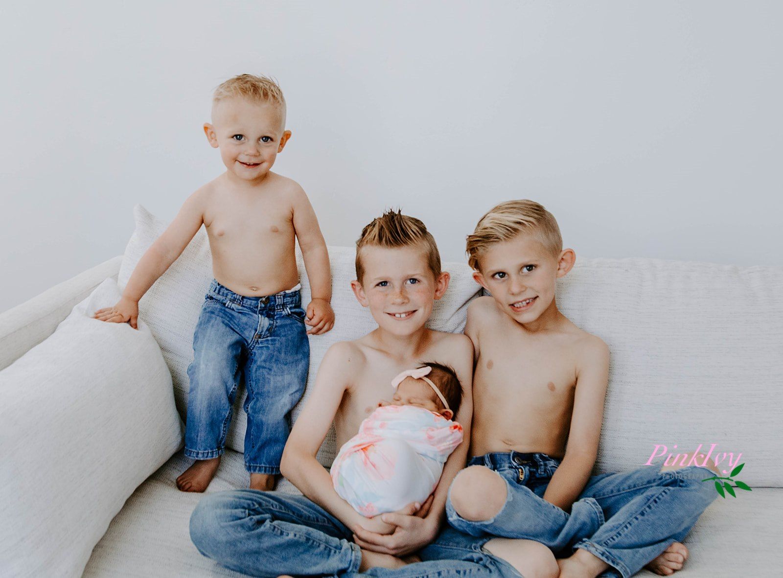 Three young children in jeans sitting together on light gray background.