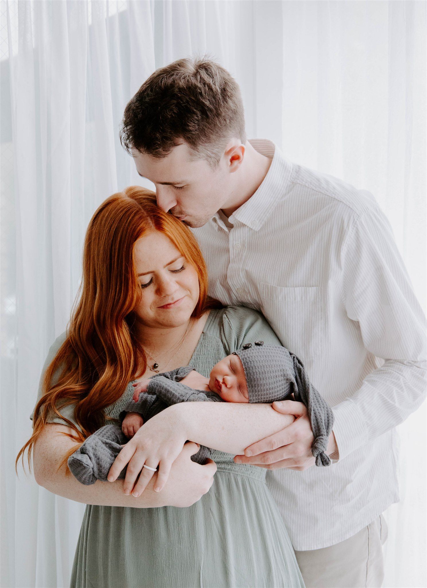 Intimate family moment with new parents in white and gray clothing by window.