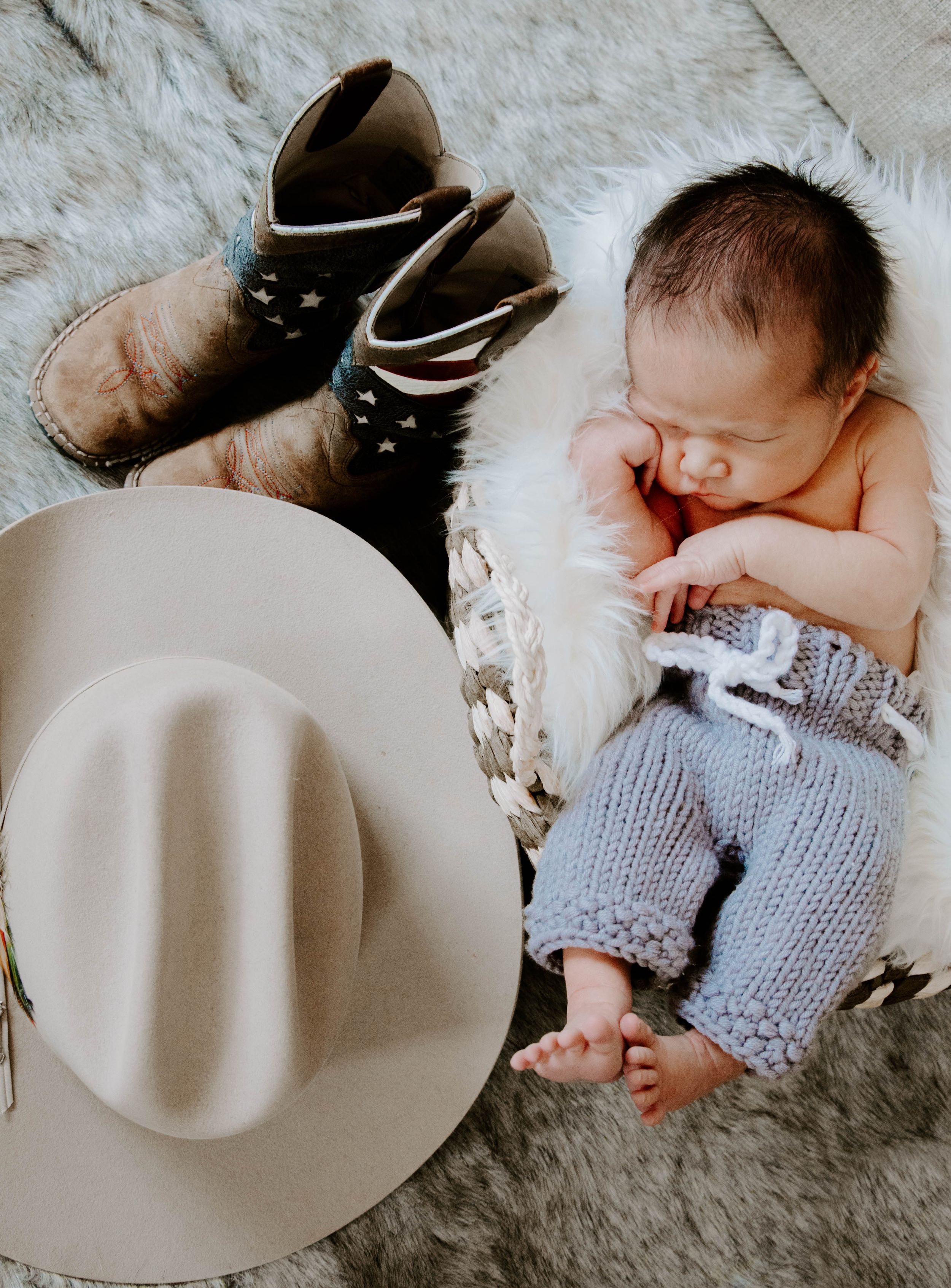 Newborn baby wearing knit pants lying on fur blanket next to brown boots and beige cowboy hat.