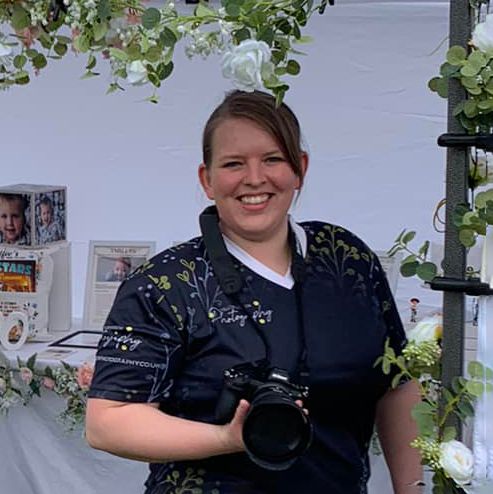 Professional photographer in navy top with camera gear stands in front of white wall with greenery.