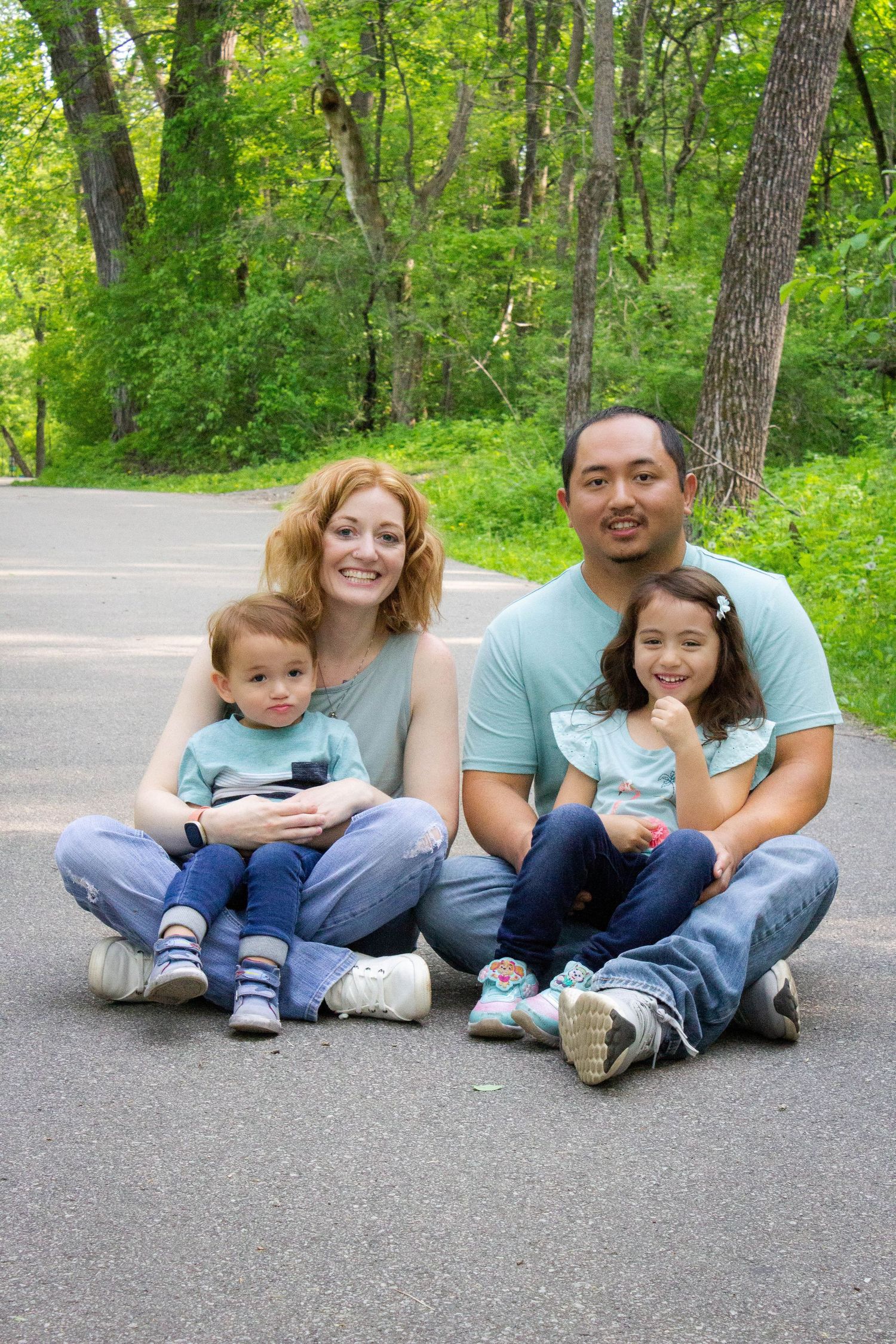 A family sits together on a paved path surrounded by green trees.