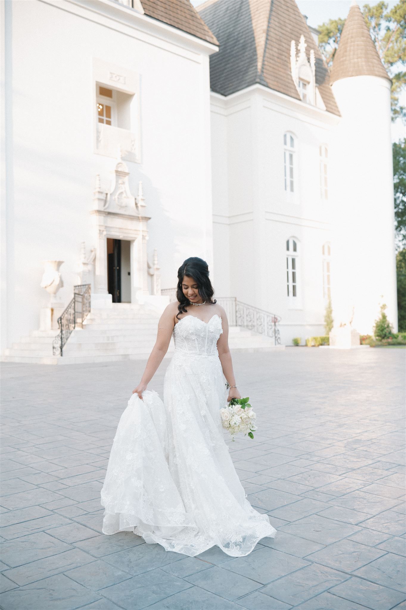 A bride in a flowing white lace wedding gown stands gracefully in front of a grand white chateau with stone details.