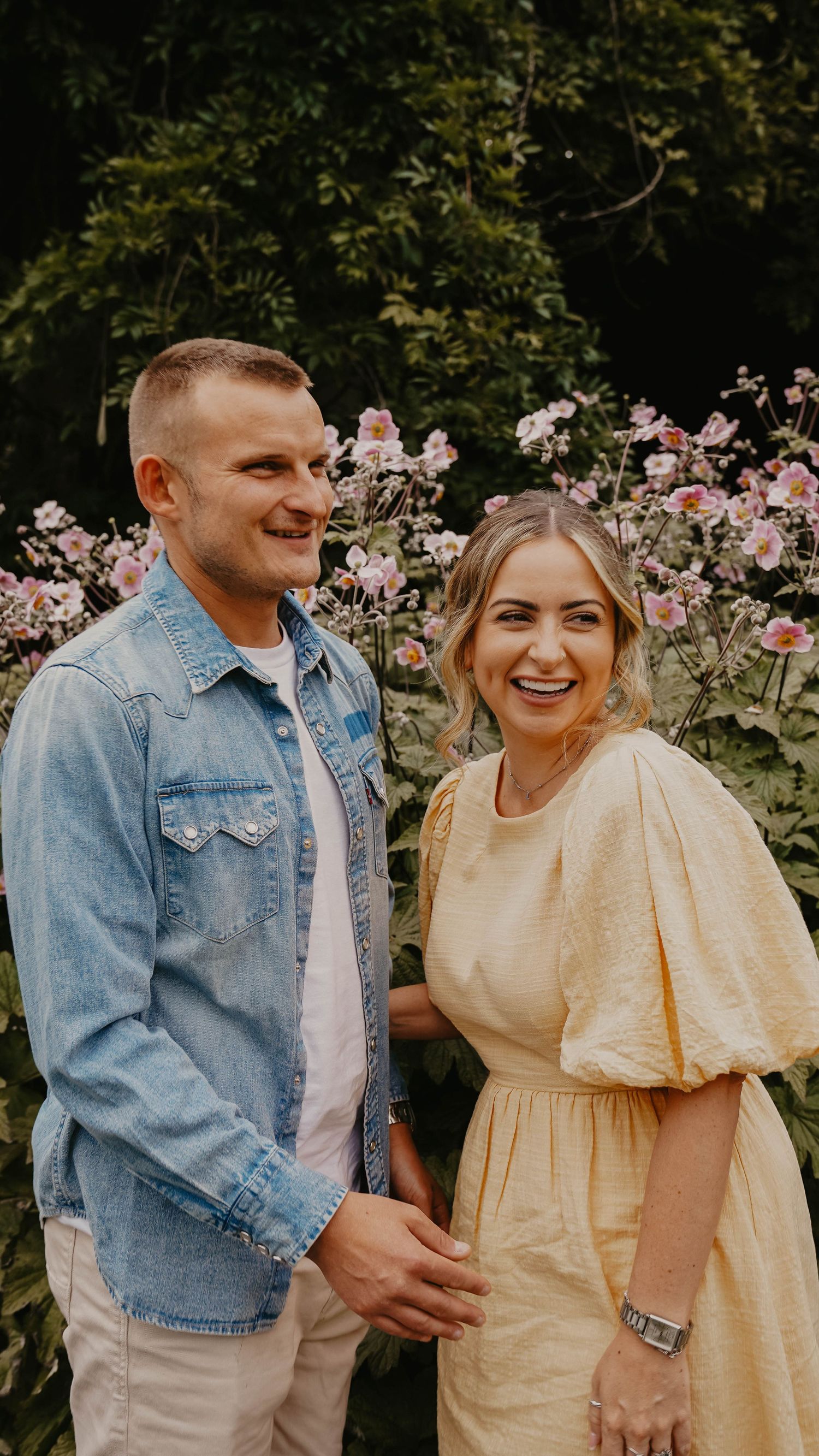 Couple in casual attire smiling together in front of blooming pink roses.