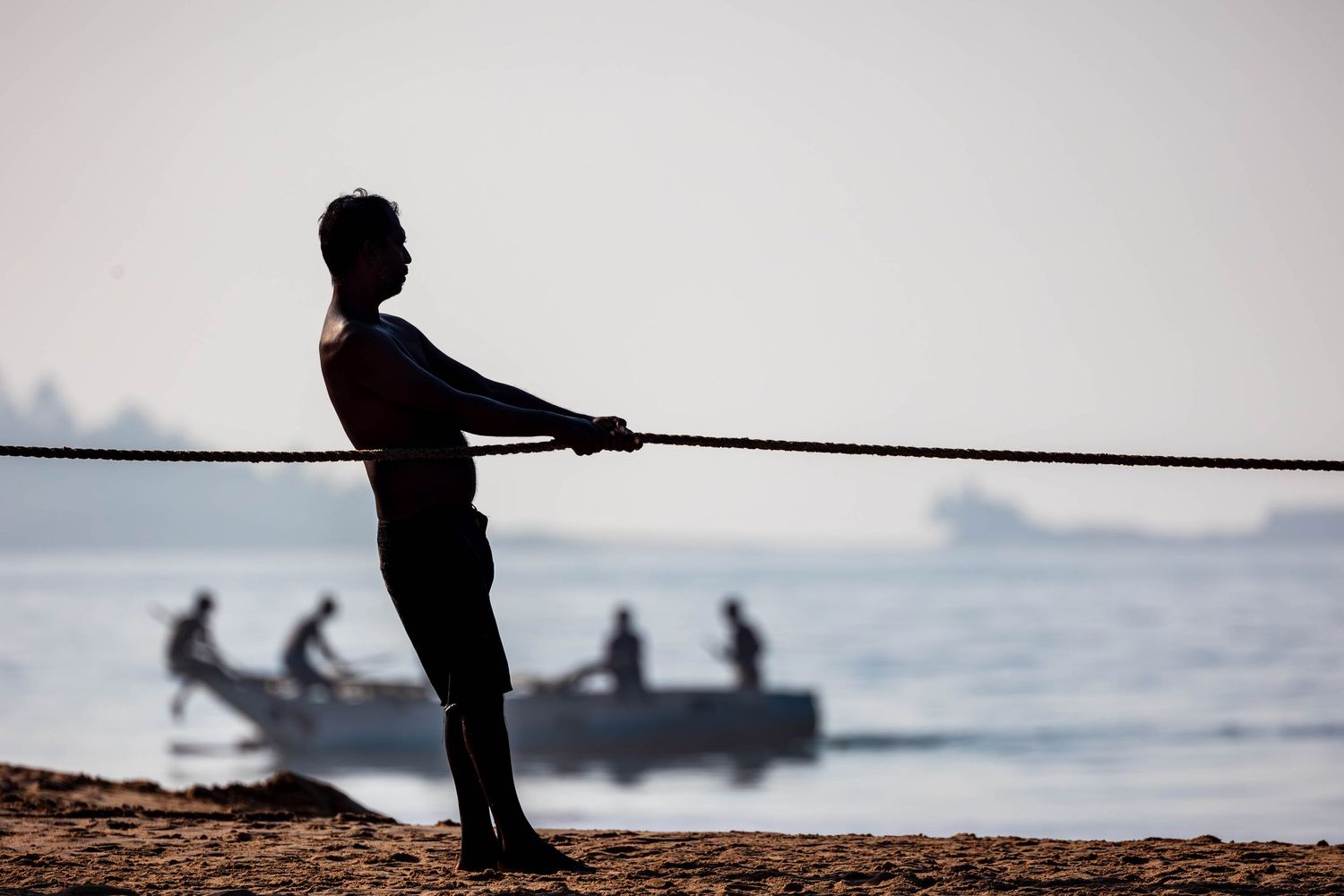 Silhouetted figure fishing from shoreline with small boat visible in misty background.