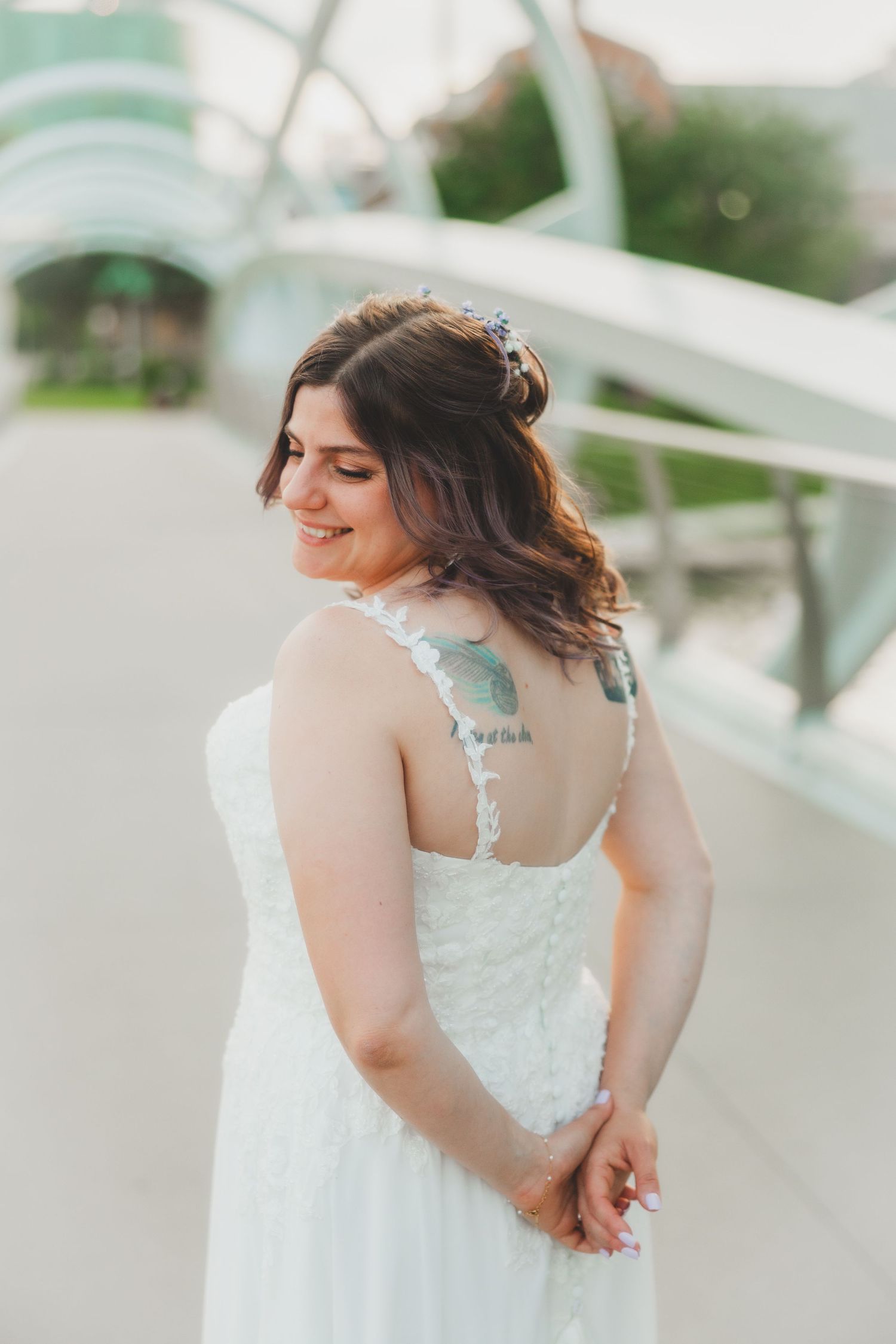 A bride in a white lace wedding dress stands on a bridge with a subtle smile and delicate hair accessories.