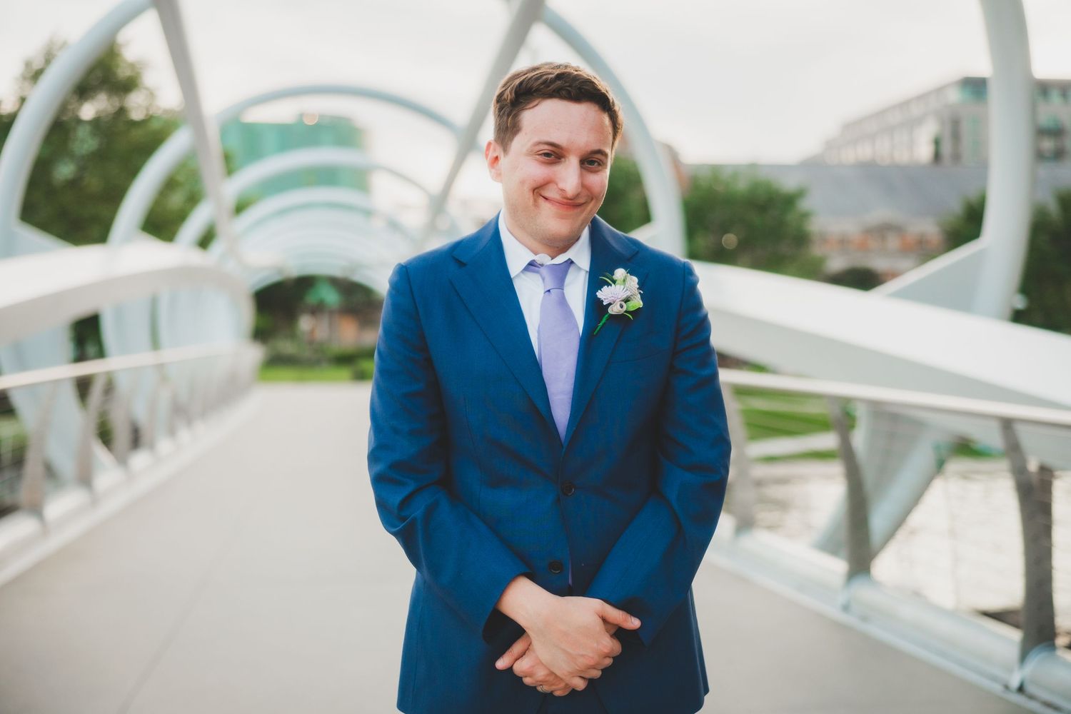 A groom in a navy blue suit and light blue tie stands on a modern bridge structure.