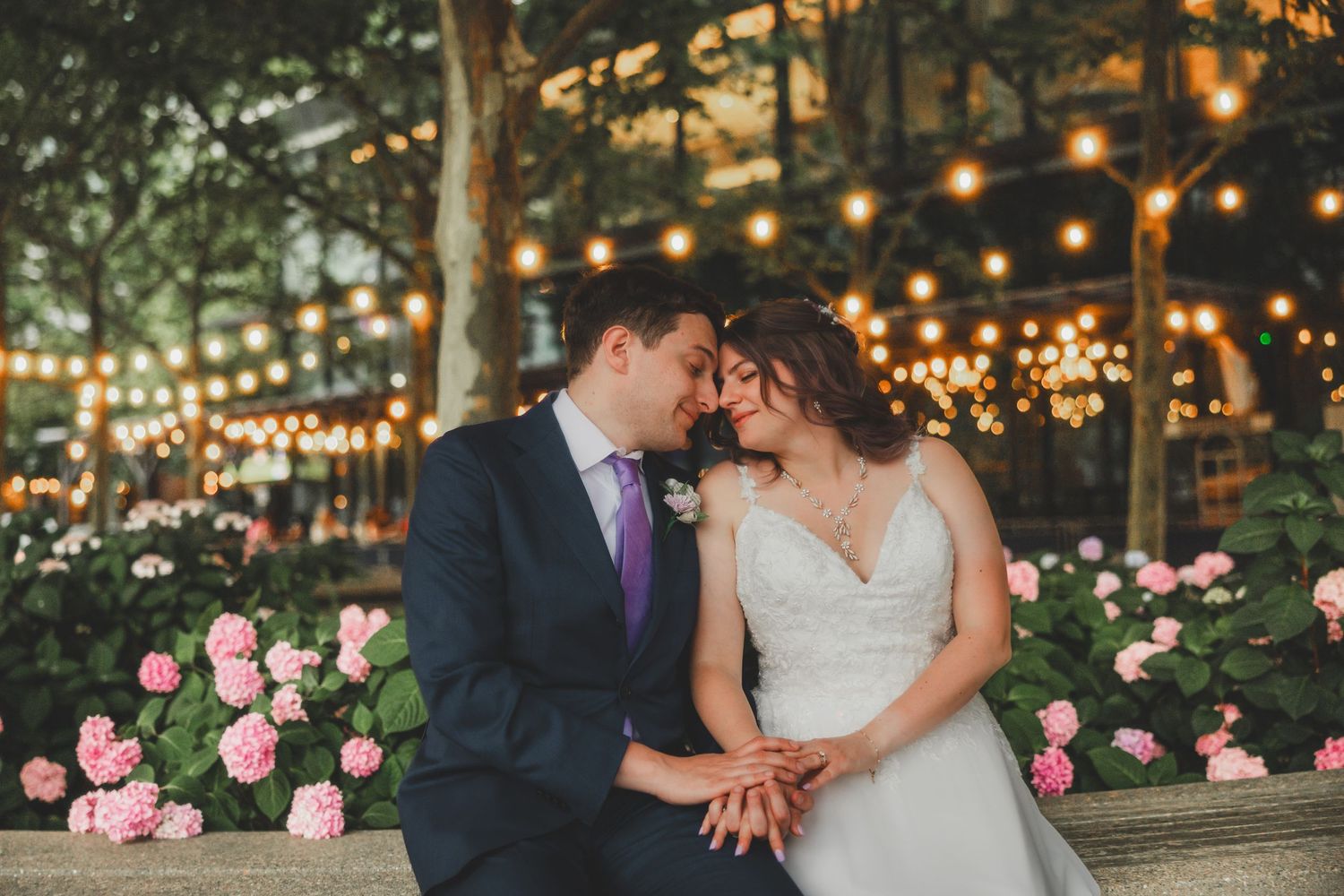 Wedding couple shares an intimate moment surrounded by pink roses and string lights in a garden setting.