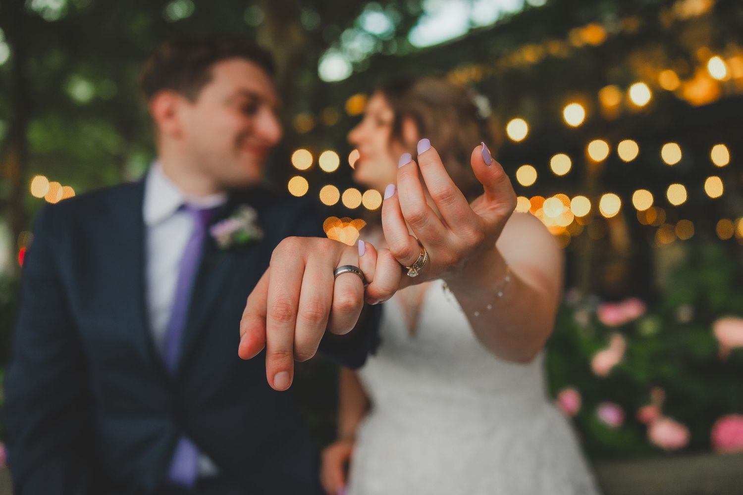 Close-up of newlyweds showing off their wedding rings with string lights glowing in background.