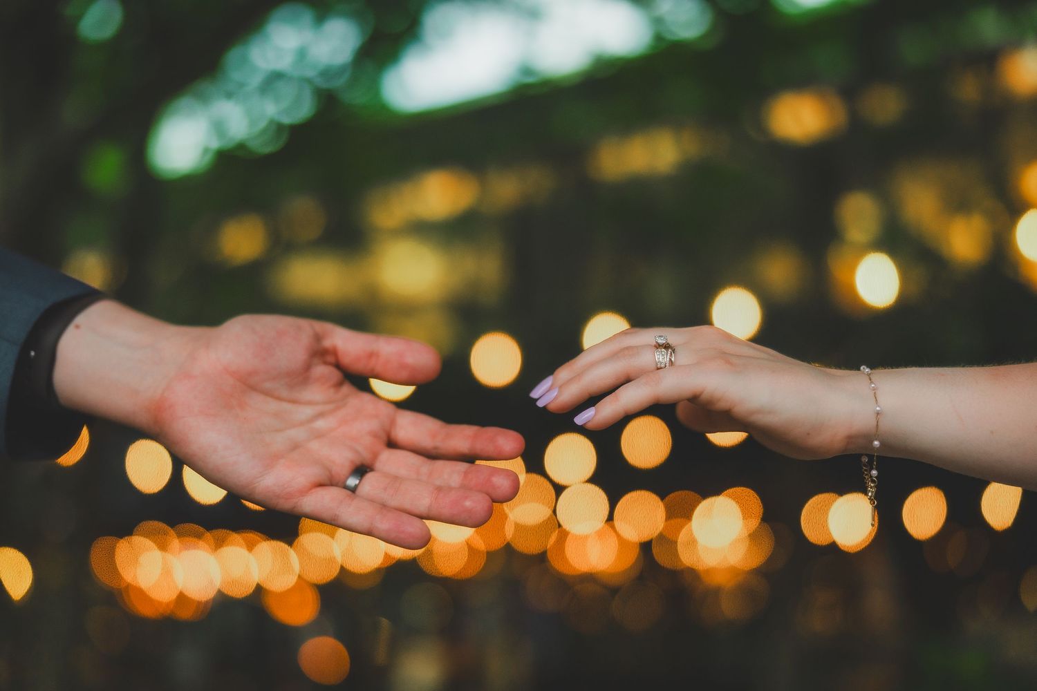 Hands reaching toward each other with wedding rings against blurred bokeh lights in evening setting.