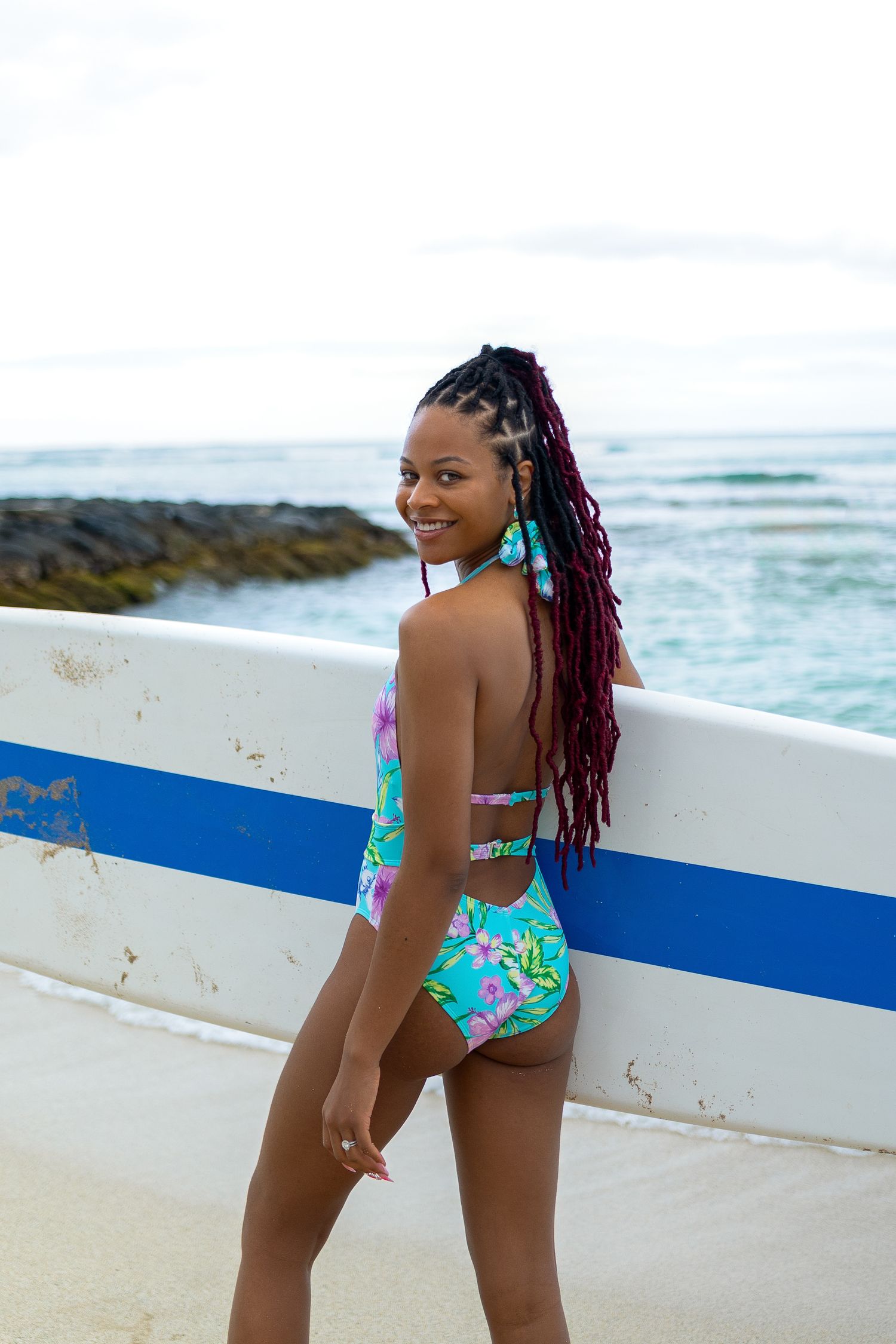 A person in a tropical print swimsuit poses next to a surfboard on a beach with ocean waves in the background.