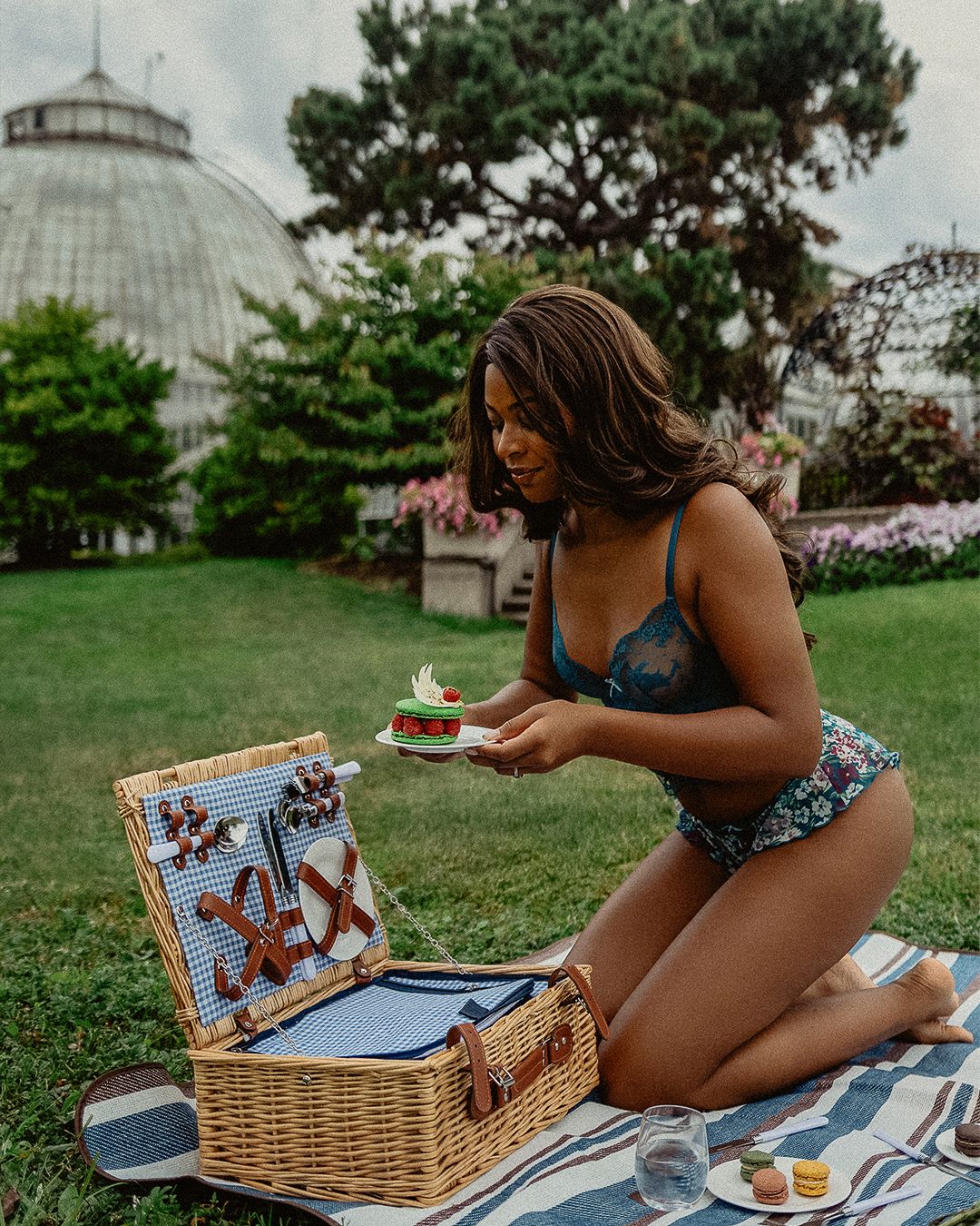 A picnic scene with a wicker basket and blanket set up on grass near a glass conservatory dome on a summer day.