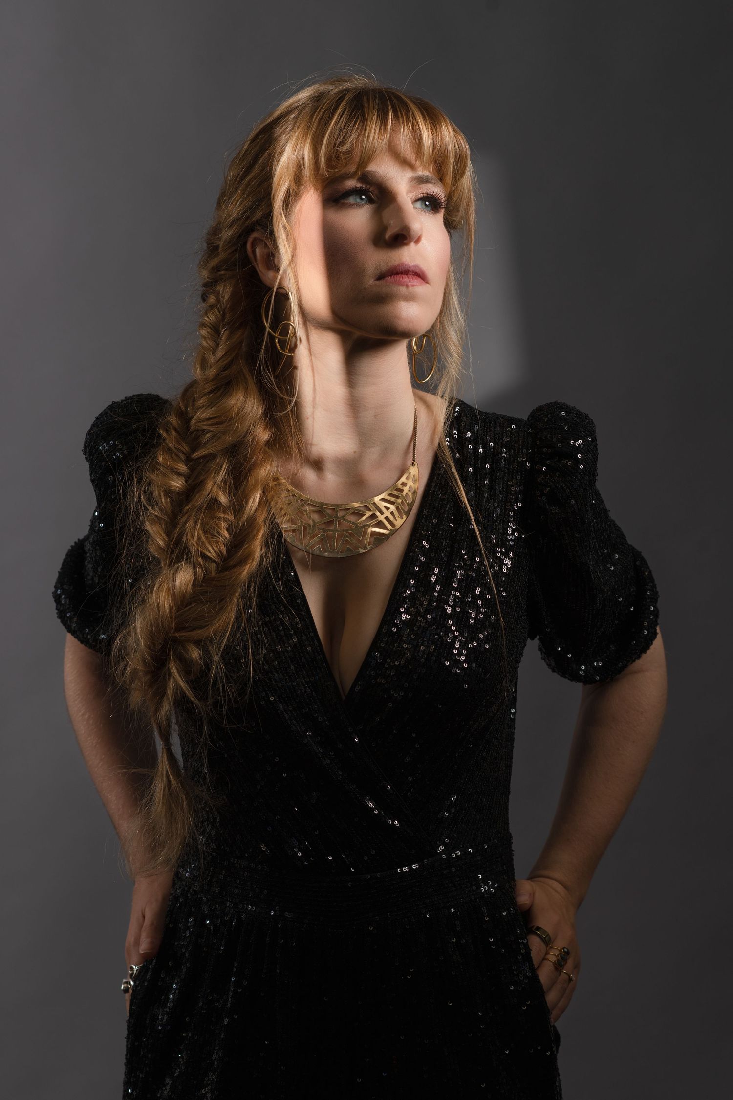 Dramatic portrait in a sparkly black evening dress with puff sleeves and chunky gold jewelry against dark background.