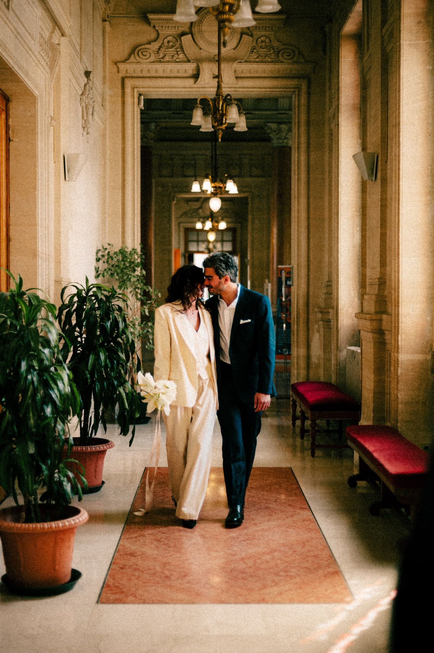 Wedding guests stand and clap in a historic venue with ornate walls and red carpet.