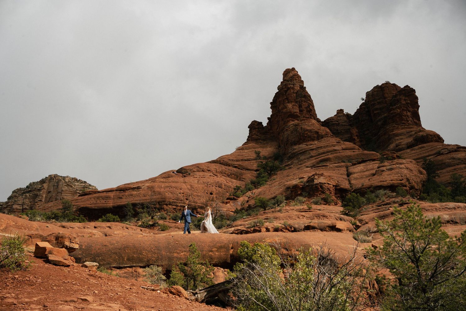 Wedding couple stands tiny against massive red rock formations in desert landscape.