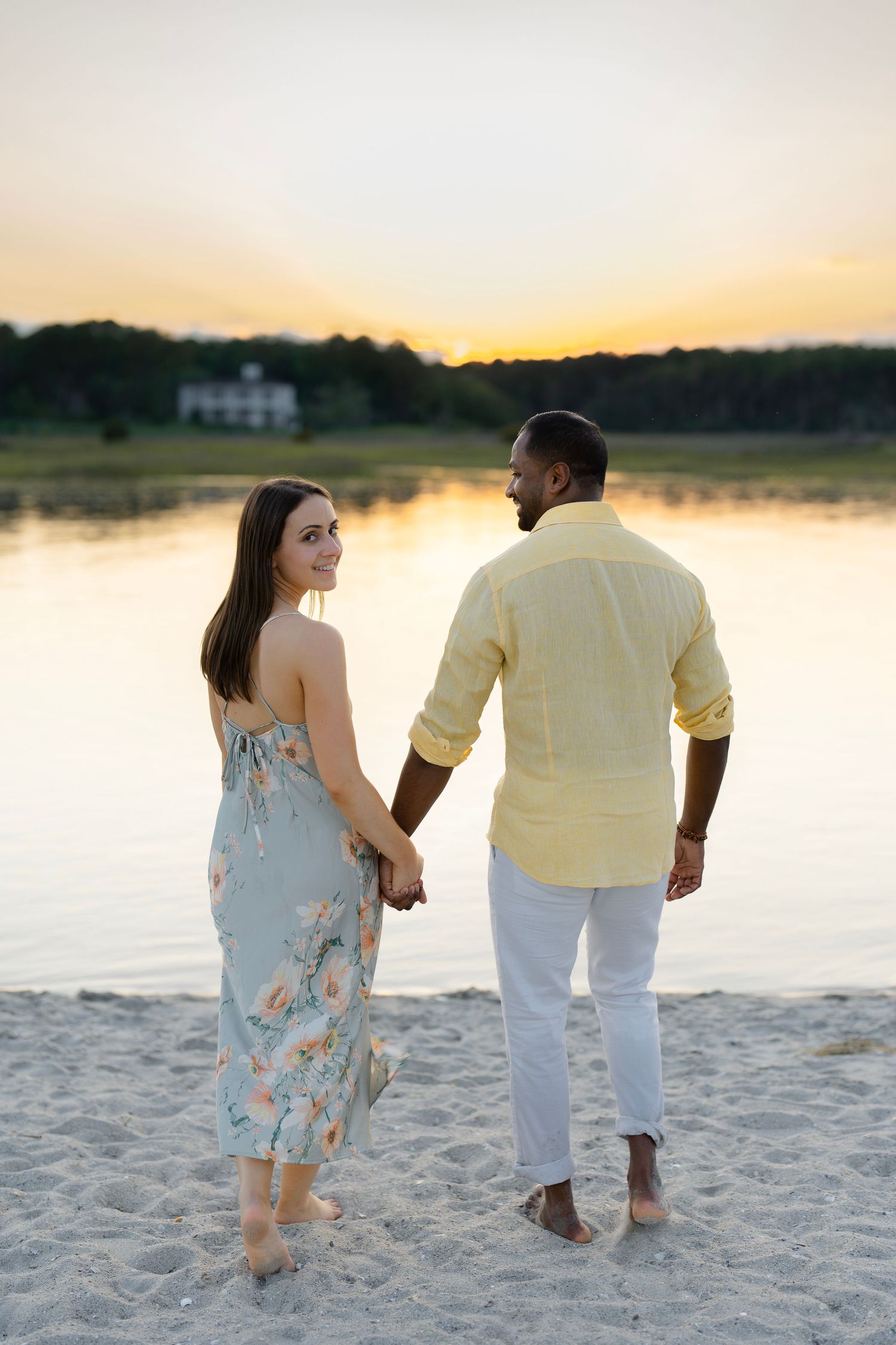 A couple walking along a sandy beach at sunset near calm waters.
