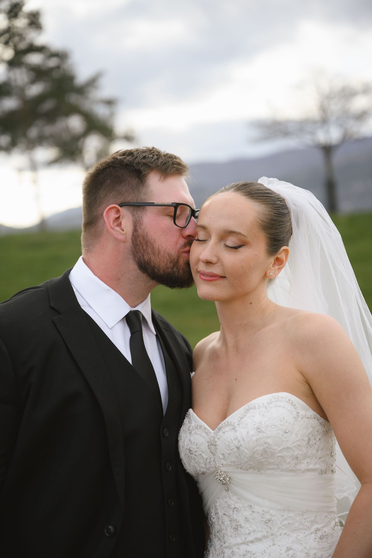Groom in black suit kissing bride on cheek outdoors, bride wearing white strapless lace gown and veil.